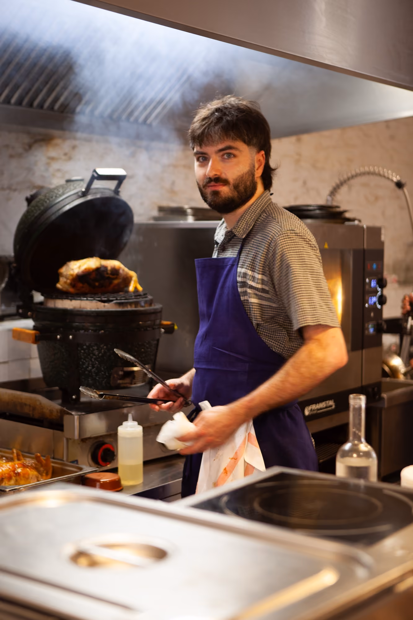 Tanguy, le chef de Midi Minuit, en tablier bleu, pinces à la main, devant le barbecue en céramique où rôtit une volaille